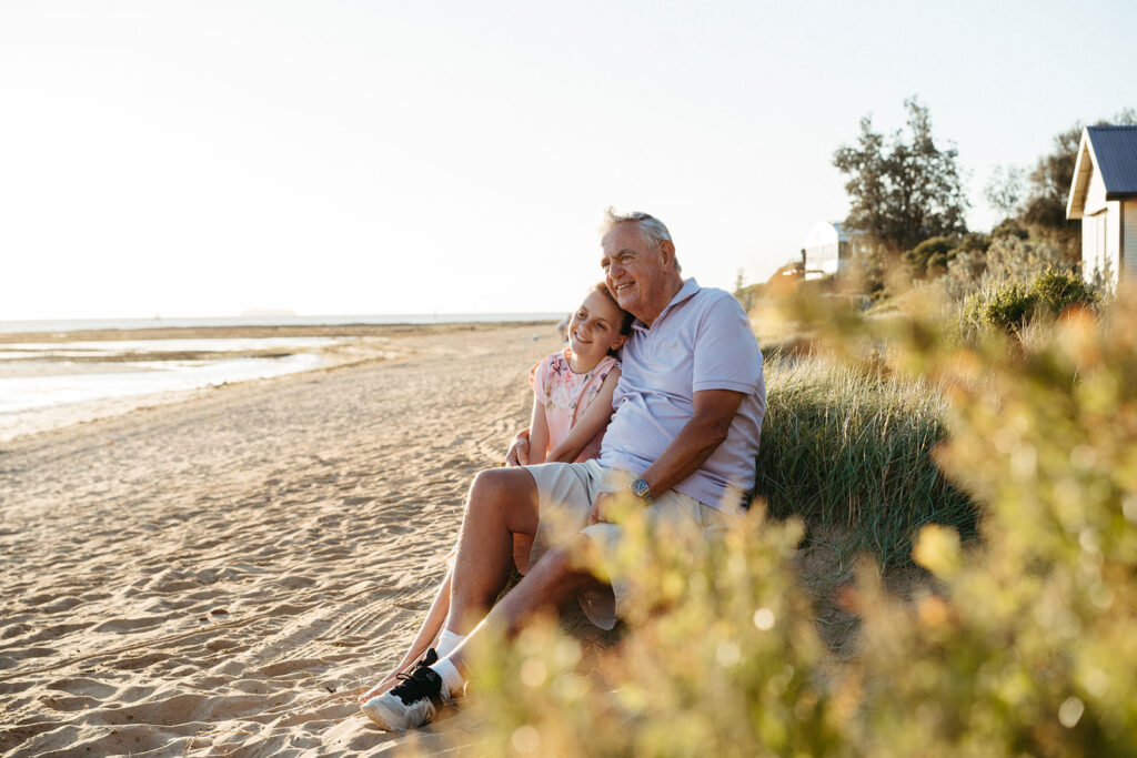 a girl sits on a sand dune at a Melbourne beach with her grandfather