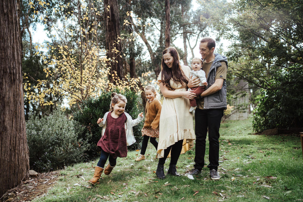 A Melbourne family standing in their green yard in autum with two young girls running around the parents