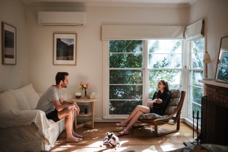 melbourne family photographer a family sits at home a father on a couch looking at his wife who is holding their baby on a seperate chair in a corner window a small dog sits at their feet in the sun