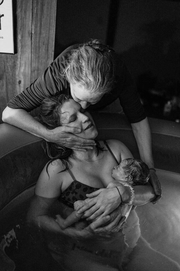 a mother kisses her adult daughter who is holding her newborn son they are sitting in a birth pool