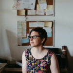 a portrait of a copywriter sitting at a desk with a wall of post it notes behind her, she is wearing glasses and a colourful dress
