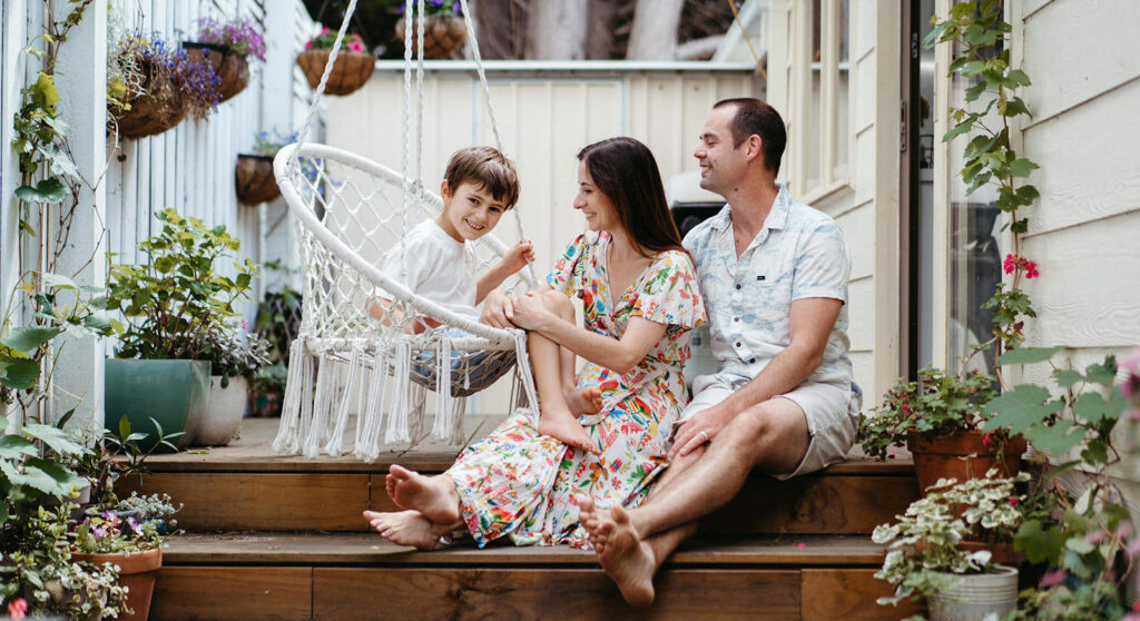a young boy sits on a macrame swing near his smiling parents on a back garden step during a family photography session