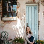 a smiling woman sits with her knees up in a doorway at a historical property for a gardeners podcast branding photoshoot
