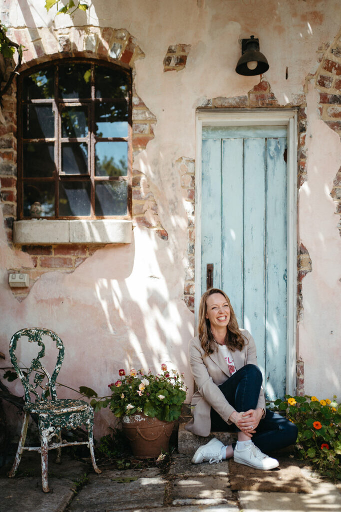 a smiling woman sits with her knees up in a doorway at a historical property for a gardeners podcast branding photoshoot