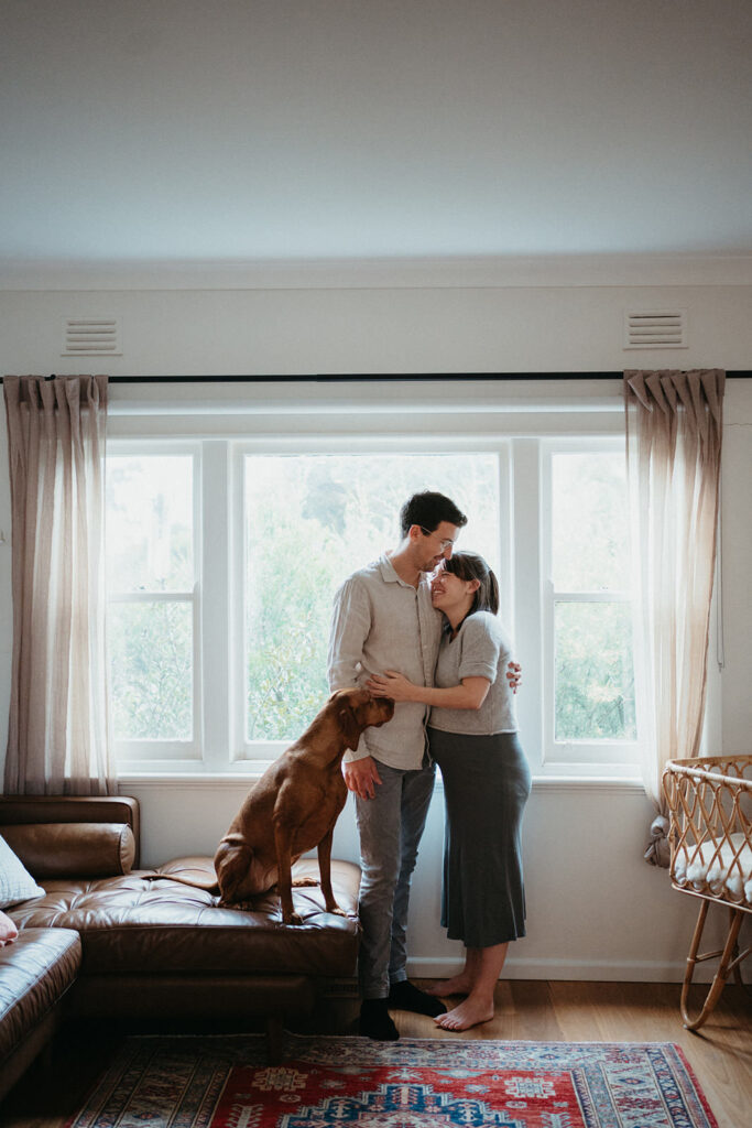 a couple stand in window, the woman is pregnant and their dog is leaning on them