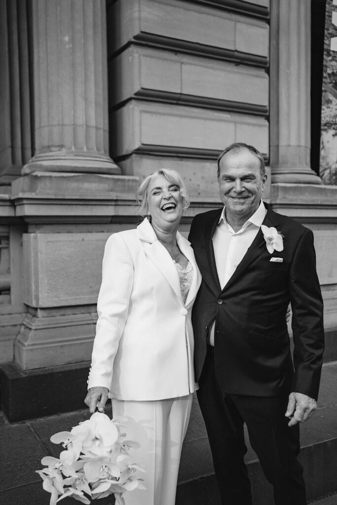 a black and white image of an older couple getting married at the registry office they are laughing and she is wearing a classic white suit